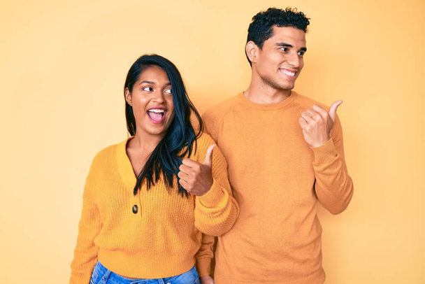Beautiful latin young couple wearing casual clothes together smiling with happy face looking and pointing to the side with thumb up.  - Photo, Image