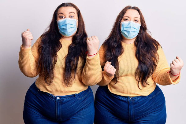 Young plus size twins wearing medical mask celebrating surprised and amazed for success with arms raised and open eyes. winner concept.  - Φωτογραφία, εικόνα