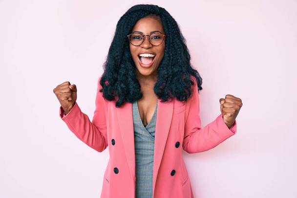 Όμορφη Αφρικανή γυναίκα που φοράει επιχειρηματικό σακάκι και γυαλιά γιορτάζει έκπληκτος και έκπληκτος για την επιτυχία με τα χέρια ψηλά και ανοιχτά μάτια. έννοια νικητή.  - Φωτογραφία, εικόνα