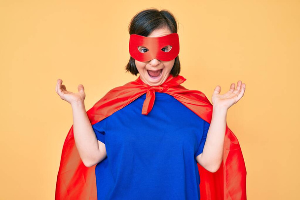 Femme brune avec le syndrome du duvet portant costume de super héros célébrant la victoire avec sourire heureux et expression gagnante avec les mains levées  - Photo, image