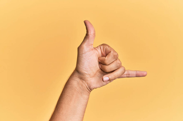 Bras et main de l'homme caucasien sur fond jaune isolé geste hawaïen shaka salutation geste, téléphone et symbole de communication  - Photo, image
