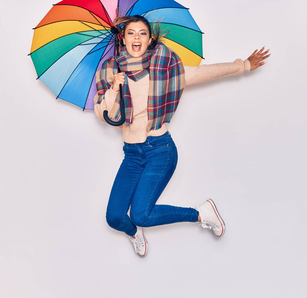 Young beautiful girl wearing winter clothes and scarf smiling happy. Jumping with smile on face holding colorful umbrella over isolated white background - Photo, Image