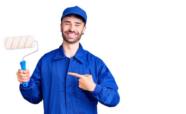 Young hispanic man wearing painter uniform holding roller smiling happy pointing with hand and finger  - Foto, Bild