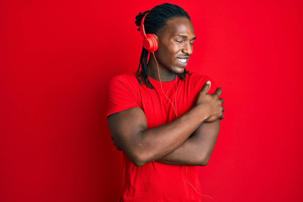 ヘッドフォンを使用して音楽を聞いているアフリカ系アメリカ人の男性は、自分自身を抱擁し、幸せと肯定的で、自信を笑っています。自己愛と自己ケア  - 写真・画像