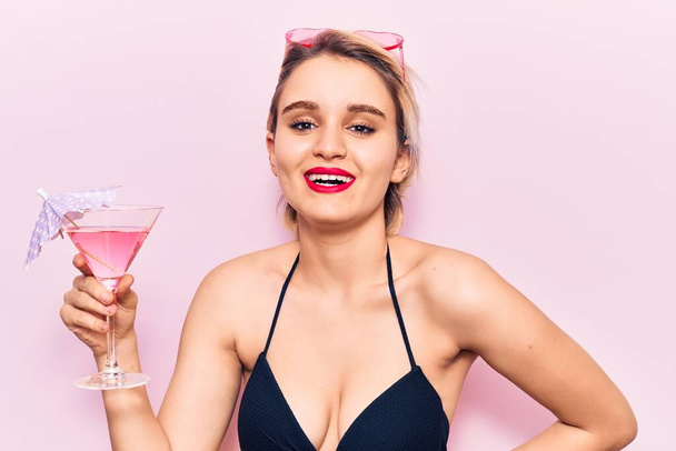 Joven hermosa mujer rubia con bikini bebiendo cóctel mirando positiva y feliz de pie y sonriendo con una sonrisa confiada mostrando los dientes  - Foto, Imagen