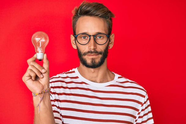 Handsome caucasian man with beard holding lightbulb for inspiration and idea thinking attitude and sober expression looking self confident  - Photo, Image