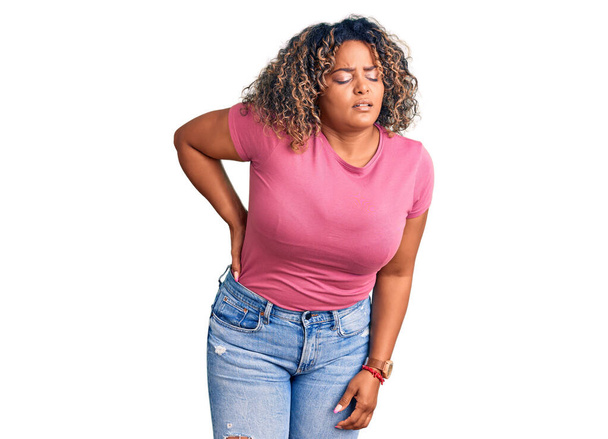 Młoda Afroamerykanka plus size kobieta ubrana w luźne ubrania cierpiąca na ból pleców, dotykająca pleców dłonią, ból mięśni  - Zdjęcie, obraz
