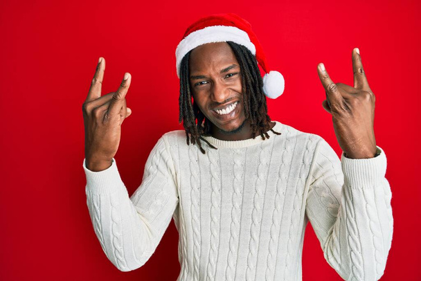 Αφροαμερικάνος με κοτσίδες φοράει χριστουγεννιάτικο καπέλο φωνάζοντας με τρελή έκφραση κάνει ροκ σύμβολο με τα χέρια ψηλά. αστέρι της μουσικής. βαριά μουσική έννοια.  - Φωτογραφία, εικόνα