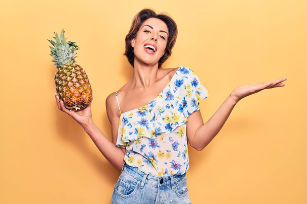 Jeune belle femme tenant ananas célébrant la réalisation avec sourire heureux et expression gagnante avec la main levée  - Photo, image