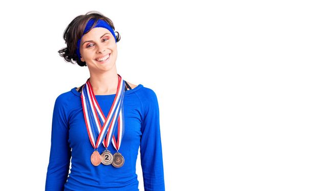 Красива молода жінка з коротким волоссям, одягнена в медалі-переможниці, виглядає позитивно і щасливо стоячи і посміхаючись з впевненою посмішкою, показуючи зуби
  - Фото, зображення