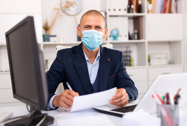 Office worker in a protective mask works behind laptop - Foto, afbeelding