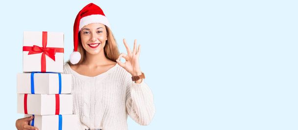 Bella giovane donna indossa il cappello di Natale e tenendo in mano un regalo facendo segno ok con le dita, sorridente gesto amichevole eccellente simbolo  - Foto, immagini