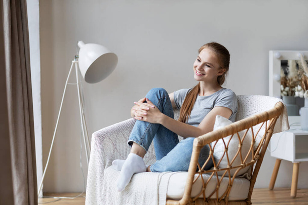Relaxed redhead girl is sitting on modern chair in light cozy room at home. Enjoying resting at home. - Photo, image