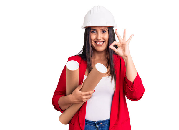 Young beautiful brunette woman wearing hardhat holding architect blueprint doing ok sign with fingers, smiling friendly gesturing excellent symbol  - 写真・画像