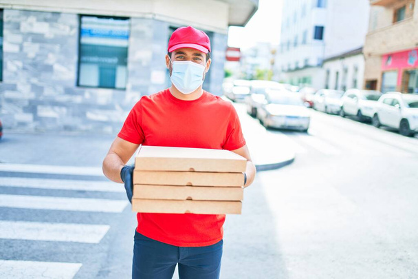 Nuori lähetti univormussa ja sepelvaltimoviruksen suojelussa. Tila toimittaa pizzalaatikoita kaupungin kadulla. - Valokuva, kuva