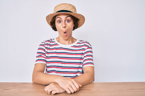 Młoda Latynoska kobieta w luźnych ubraniach i kapeluszu siedząca na stole przerażona i zszokowana zaskakującą ekspresją, strachem i podnieconą twarzą.  - Zdjęcie, obraz