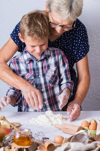 Ευτυχισμένη ώριμη γυναίκα, γιαγιά και αγόρι, εγγονός που μαγειρεύει, ζυμώνει ζύμη, πίτα, κέικ, μπισκότα. Οικογενειακή στιγμή στη ζεστή κουζίνα. Φθινόπωρο δραστηριότητα στο σπίτι. - Φωτογραφία, εικόνα