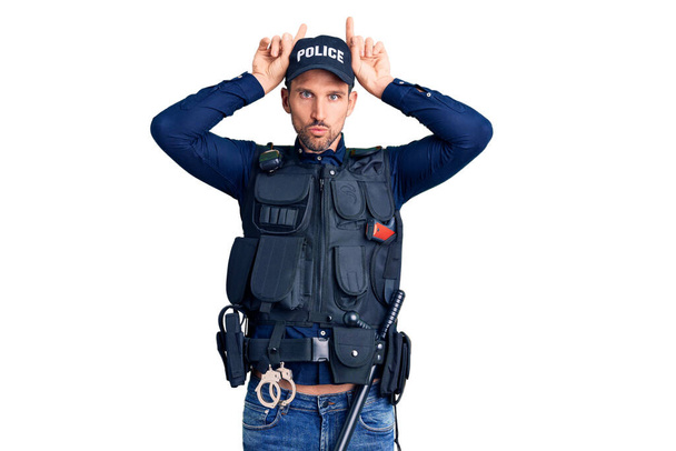 Młody przystojny mężczyzna w mundurze policyjnym robi zabawny gest z palcem nad głową jak rogi byka  - Zdjęcie, obraz