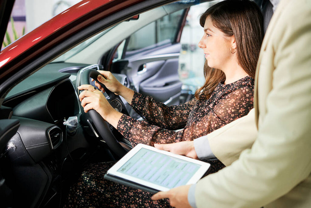 Close-up van de manager van de auto salon staan met tablet pc en suggereren aan vrouwelijke klant om haar nieuwe auto te verzekeren - Foto, afbeelding