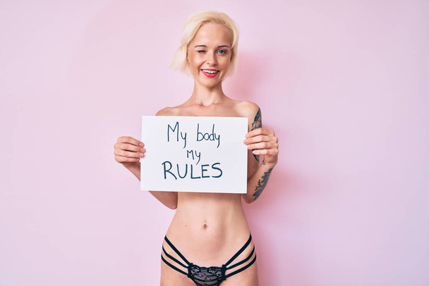 Young blonde woman with tattoo standing shirtless holding banner with my body my rules message winking looking at the camera with sexy expression, cheerful and happy face.  - Photo, Image
