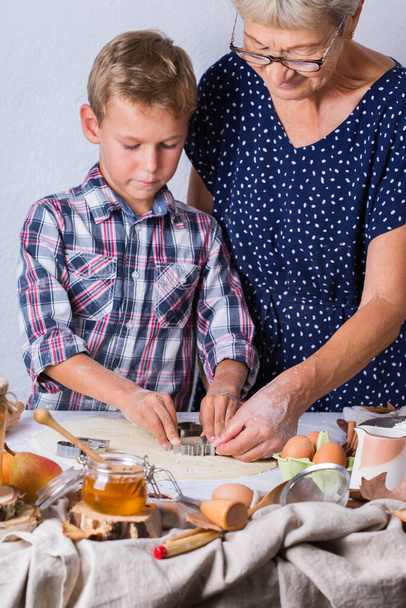 Ευτυχισμένη ώριμη γυναίκα, γιαγιά και αγόρι, εγγονός που μαγειρεύει, ζυμώνει ζύμη, πίτα, κέικ, μπισκότα. Οικογενειακή στιγμή στη ζεστή κουζίνα. Φθινόπωρο δραστηριότητα στο σπίτι. - Φωτογραφία, εικόνα