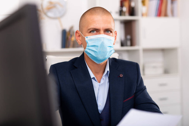 Office worker in a protective mask works behind laptop - Фото, изображение