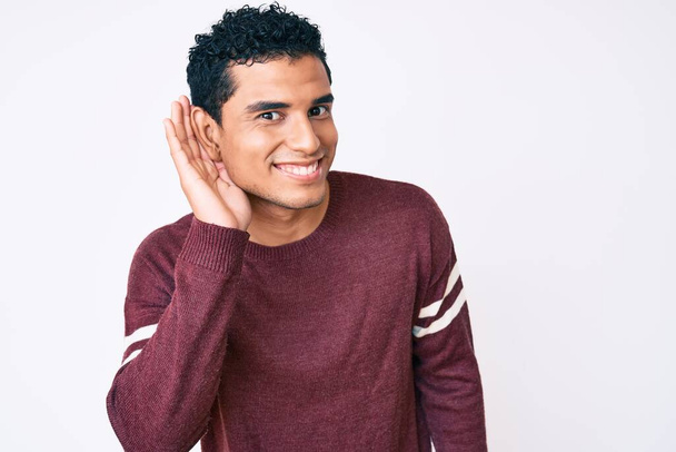 Young handsome hispanic man over white background smiling with hand over ear listening and hearing to rumor or gossip. deafness concept.  - Photo, Image