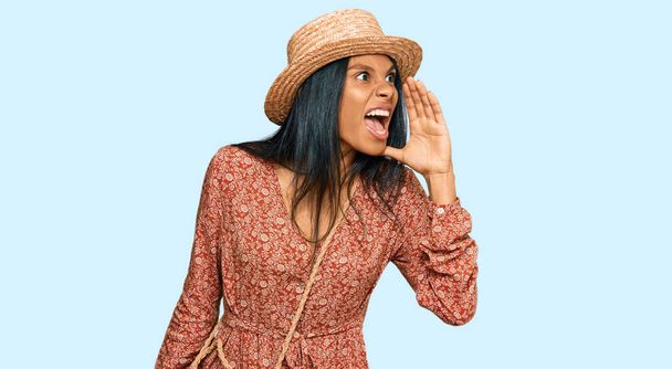 Mladá Afroameričanka v letním klobouku křičela a křičela na stranu s rukou na ústech. komunikační koncept.  - Fotografie, Obrázek