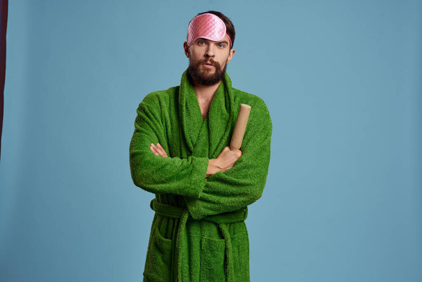 un hombre con una máscara de sueño rosa sostiene un rodillo en su mano y un modelo de emociones de fondo azul bata verde - Foto, Imagen