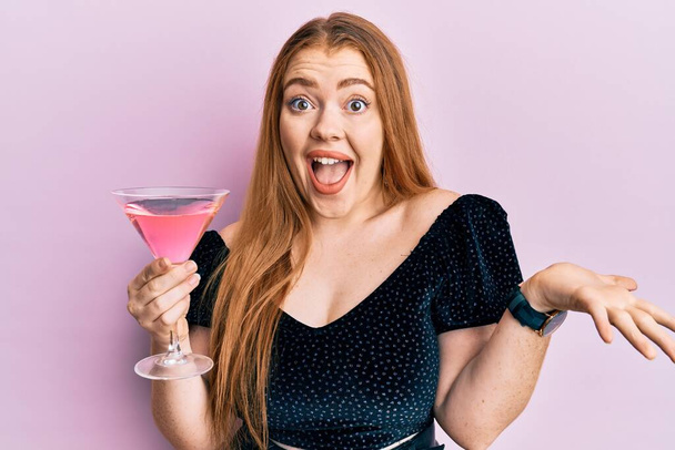Young beautiful redhead woman drinking tropical cocktail with alcohol celebrating achievement with happy smile and winner expression with raised hand  - Foto, Imagem