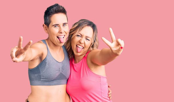 Couple of women wearing sportswear smiling with tongue out showing fingers of both hands doing victory sign. number two.  - Photo, Image