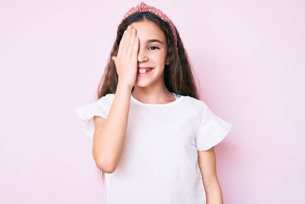 Günlük kıyafetler giyen tatlı İspanyol kız çocuğu ve bir gözünü eliyle kapatan diyabet, yüzünde kendine güvenen bir gülümseme ve sürpriz bir duygu..  - Fotoğraf, Görsel