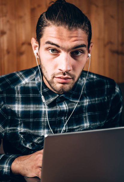 Een knappe jonge freelancer in een blauw shirt en witte hoofdtelefoon werkt thuis op een zilveren laptop. Bediening op afstand tijdens de quarantaine, om de tekst af te drukken en belangrijke zaken te doen. - Foto, afbeelding