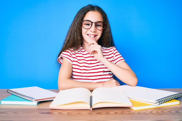 Χαριτωμένο ισπανόφωνο κορίτσι που σπουδάζει για τις σχολικές εξετάσεις κάθεται στο τραπέζι χαμογελώντας κοιτάζοντας αυτοπεποίθηση στην κάμερα με σταυρωμένα χέρια και το χέρι στο πηγούνι. θετική σκέψη.  - Φωτογραφία, εικόνα
