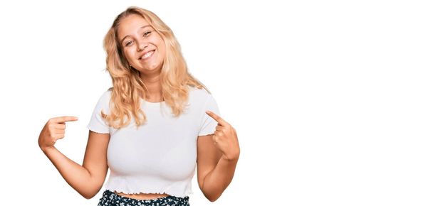Junges blondes Mädchen in lässigem weißem T-Shirt sieht selbstbewusst aus mit einem Lächeln im Gesicht und zeigt stolz und glücklich mit den Fingern.  - Foto, Bild