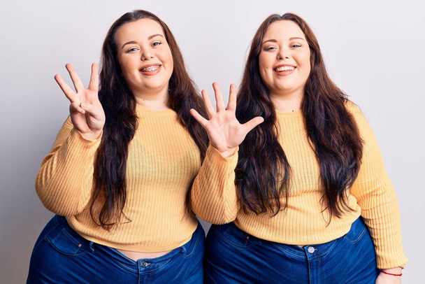 Junge Plus-Size-Zwillinge in lässiger Kleidung zeigen mit Finger Nummer acht nach oben und lächeln dabei selbstbewusst und glücklich.  - Foto, Bild