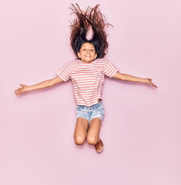 Imádnivaló spanyol kislány alkalmi ruhát visel, boldogan mosolyog. Ugrás mosollyal az arcon és a karok nyitott elszigetelt rózsaszín háttér - Fotó, kép