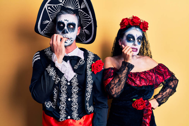 Młoda para ubrana w meksykański dzień martwego kostiumu na żółto wyglądająca na zestresowaną i zdenerwowaną z rękami obgryzającymi paznokcie. niepokój.  - Zdjęcie, obraz