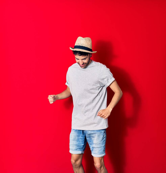 Junger gutaussehender hispanischer Mann in lässiger Kleidung und Sommermütze, der glücklich lächelt. Mit einem Lächeln im Gesicht hüpfend und mit erhobenen Fäusten über den abgetrennten roten Hintergrund feiernd. - Foto, Bild