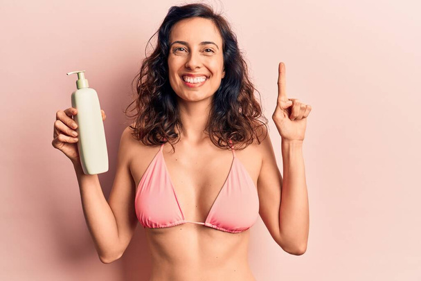 Jeune belle hispanique femme portant un bikini tenant crème solaire lotion sourire avec une idée ou une question pointant du doigt avec un visage heureux, numéro un  - Photo, image