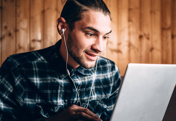A handsome young freelancer in a blue shirt and white headphones works on a silver laptop at home. Remote operation during the quarantine, to print the text and conduct important business. - Φωτογραφία, εικόνα