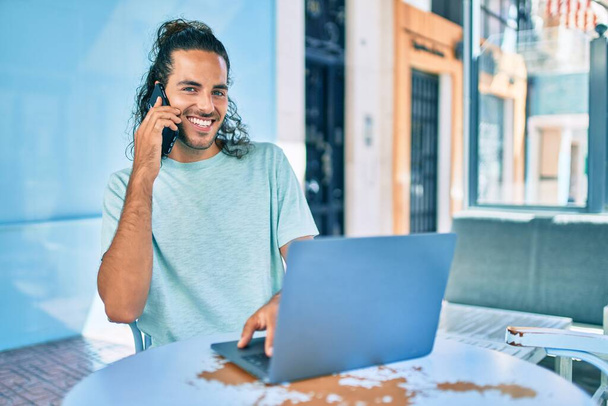 Νεαρός Ισπανός χαμογελάει χαρούμενος δουλεύοντας με λάπτοπ και μιλώντας στο κινητό του στην ταράτσα της καφετέριας.. - Φωτογραφία, εικόνα