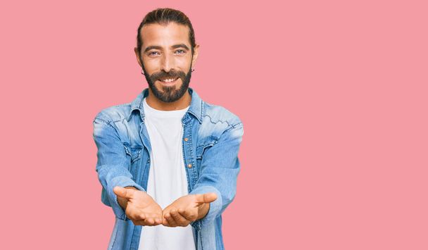 Attractive man with long hair and beard wearing casual denim jacket smiling with hands palms together receiving or giving gesture. hold and protection  - Foto, Bild