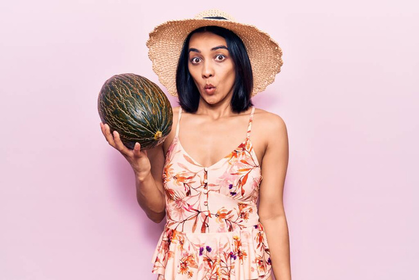 jong mooi latijn vrouw dragen zomer hoed houden meloen bang en verbaasd met open mond voor verrassing, ongeloof gezicht  - Foto, afbeelding