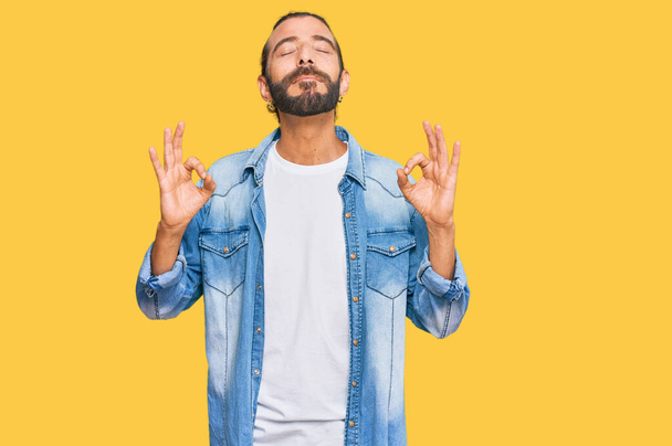Attractive man with long hair and beard wearing casual denim jacket relaxed and smiling with eyes closed doing meditation gesture with fingers. yoga concept.  - Photo, Image