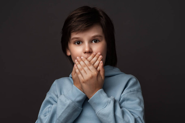 I'm afraid. Image of scared boy 10-12 years old in casual clothes covering his mouth with hands. Fright, phobia, panic attack, horror and facial expression concept. Studio shot, gray background - Foto, immagini