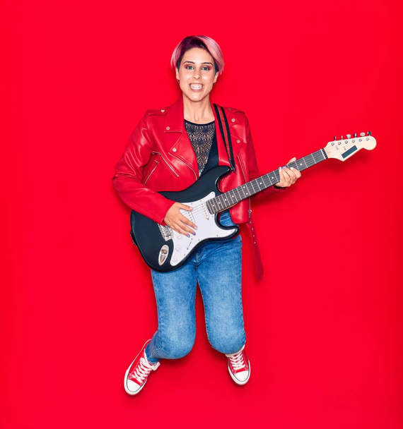 Genç, güzel gitarist, pembe kısa saçlı, gitar çalan mutlu bir kadın. İzole edilmiş kırmızı arka planda yüzünde gülümsemeyle zıplıyor - Fotoğraf, Görsel