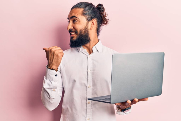 Genç Arap adam dizüstü bilgisayarını tutuyor başparmağıyla yan tarafa bakıyor mutlu bir şekilde ağzı açık gülümsüyor.  - Fotoğraf, Görsel