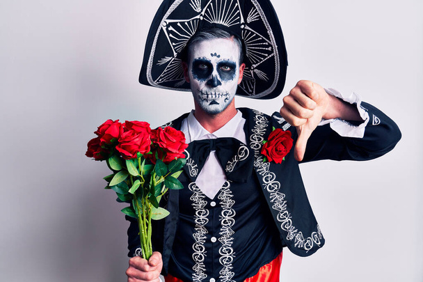 Junger Mann im mexikanischen Totenkostüm mit Blumen und wütendem Gesicht, negatives Zeichen für Abneigung mit gesenktem Daumen, Ablehnungskonzept  - Foto, Bild