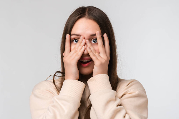 Young beautiful brunette woman covering face and eyes with hand, looking through fingers, wearing stylish beige hoodie. Human emotions, facial expression concept - Фото, изображение
